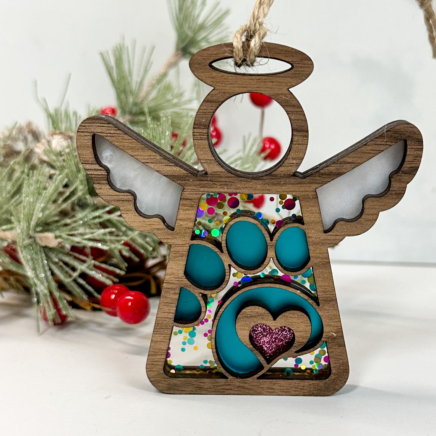 Wooden stained glass looking paw print angel ornament sitting on a white background with pine and berry sprigs
