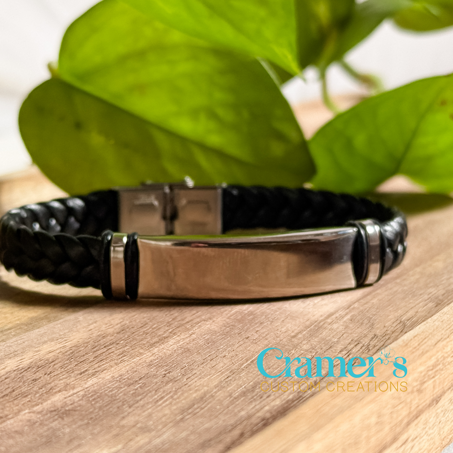 A black braided cord bracelet with a silver stainless steel metal bar, displayed on a wooden surface with a green leafy background.