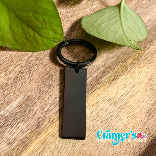 A black rectangle keychain with a keyring on one end and a leaf in the background.