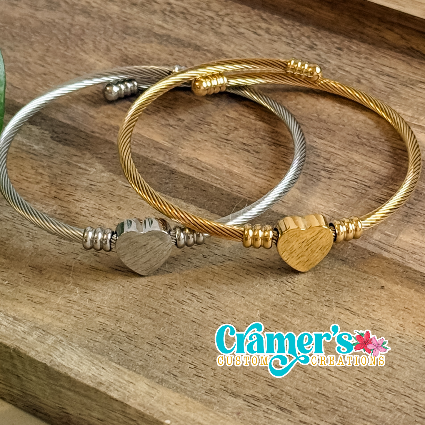 Two bracelets, one gold and one silver, with heart-shaped beads on a wooden surface, featuring the 'Cramer's Custom Creations' logo.