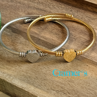 Two bracelets, one gold and one silver, with heart-shaped beads on a wooden surface, featuring the 'Cramer's Custom Creations' logo.