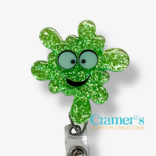 Green glittery germ-shaped badge holder with a smiley face on a white background, featuring the brand 'Cramer's Custom Creations'.