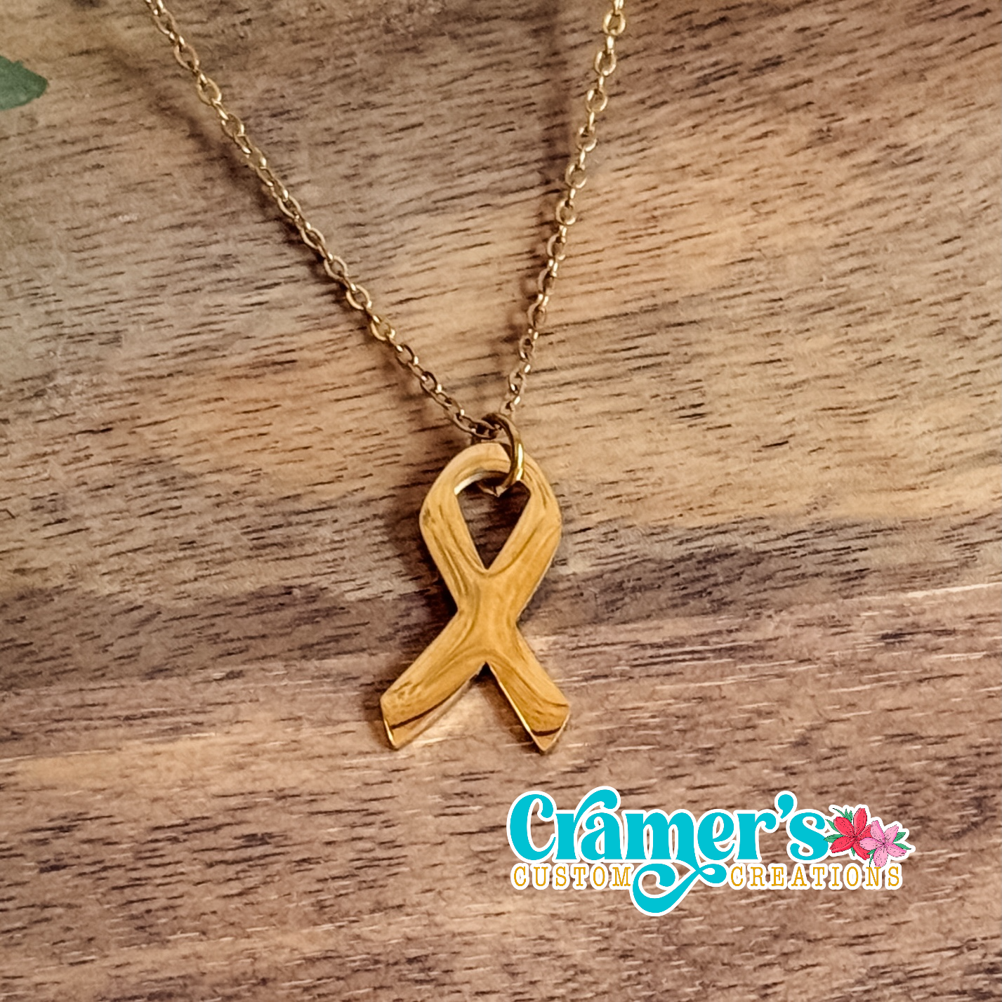 Gold ribbon necklace on a wooden surface with 'Cramer's Custom Creations' logo.