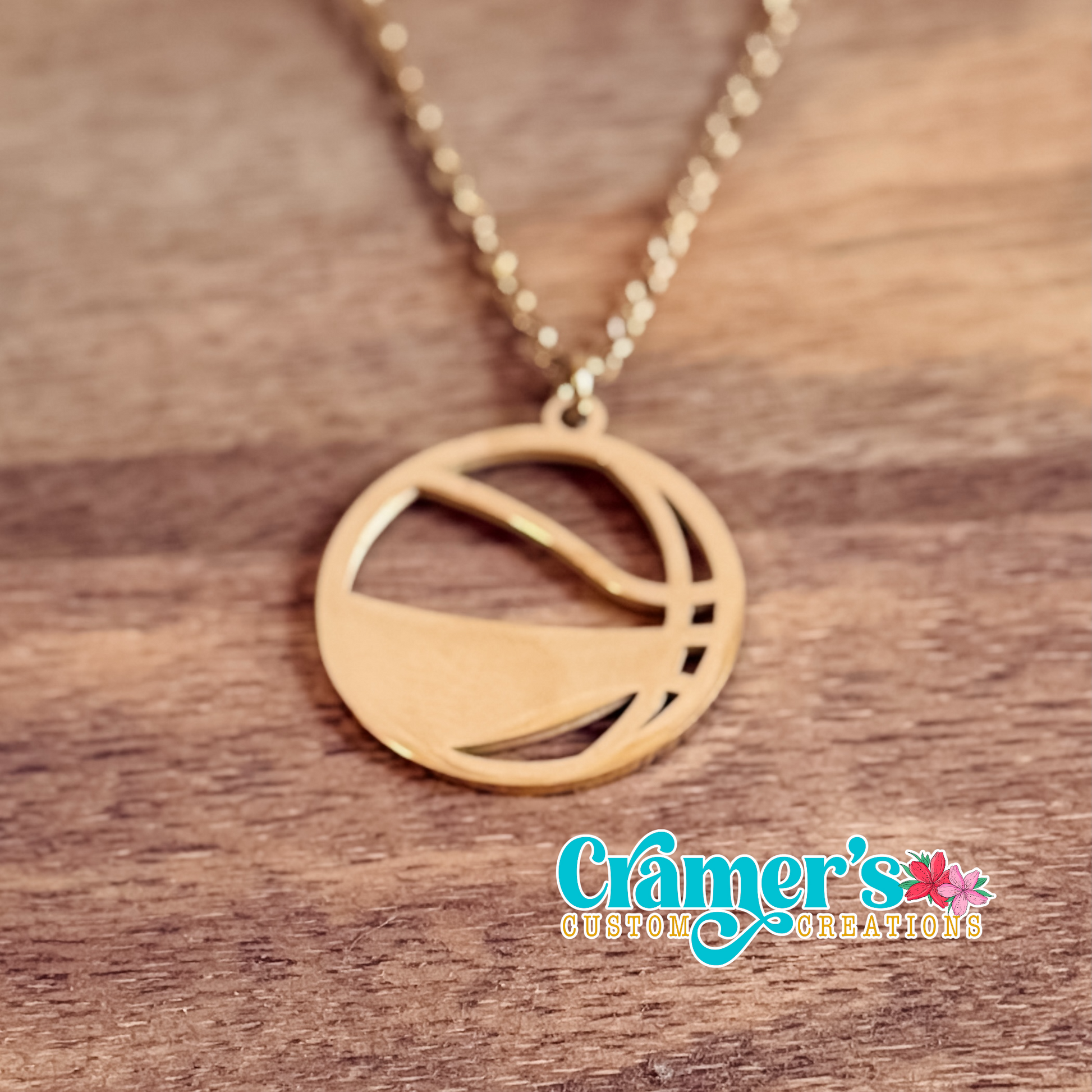 Gold necklace with a basketball pendant on a wooden surface, featuring Cramer's Custom Creations logo.