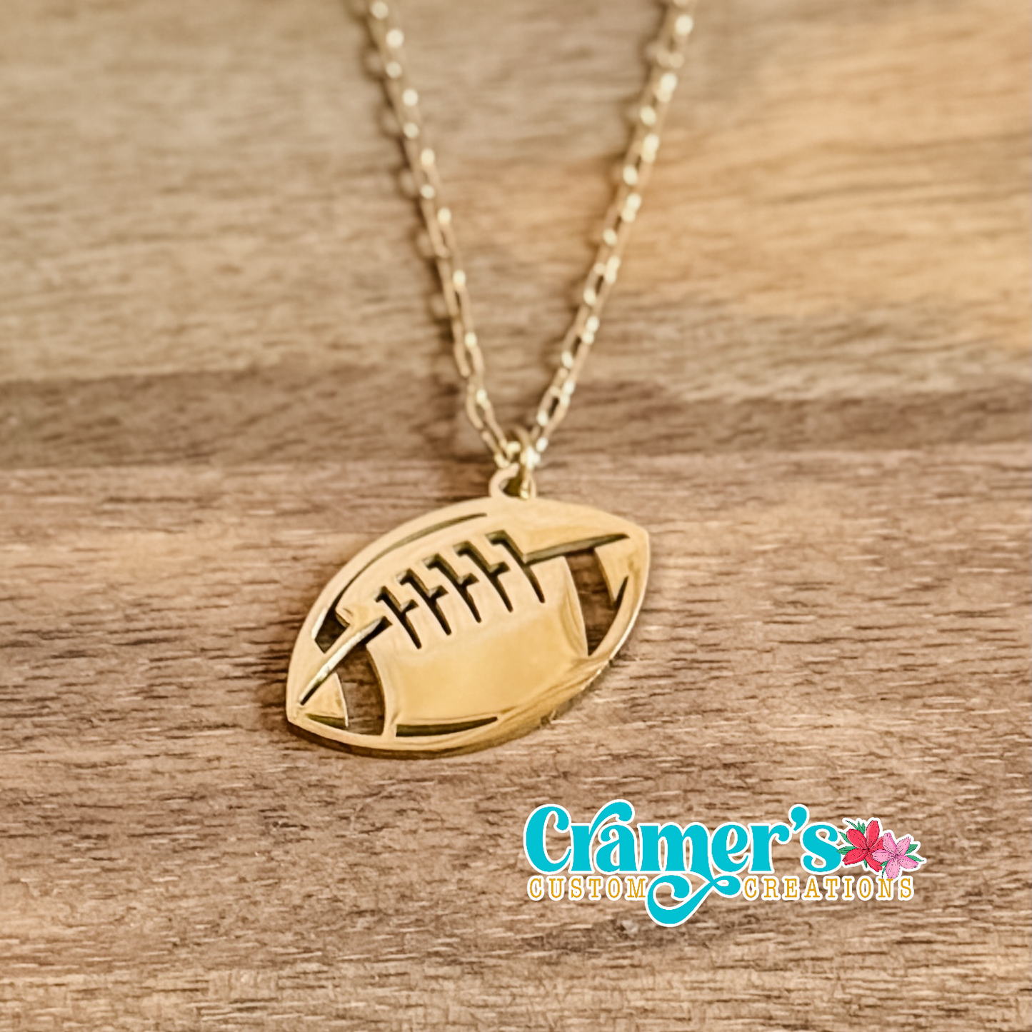 Gold football-shaped pendant necklace on a wooden surface with 'Cramer's Custom Creations' branding.