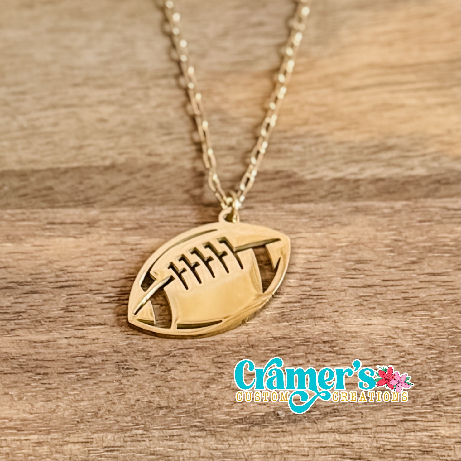 Gold football-shaped pendant necklace on a wooden surface with 'Cramer's Custom Creations' branding.