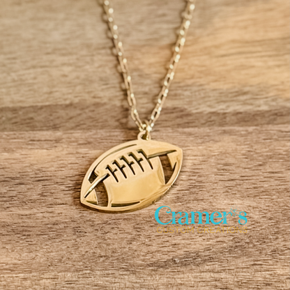 Gold football-shaped pendant necklace on a wooden surface with 'Cramer's Custom Creations' branding.