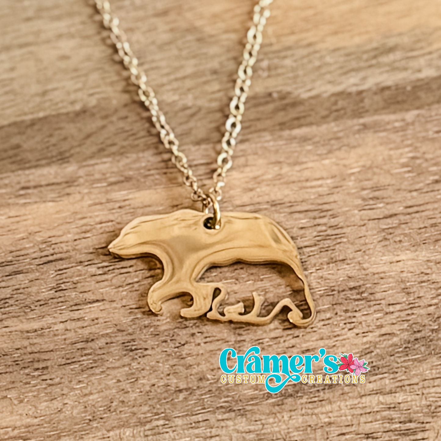 Gold necklace with a bear pendant on a wooden surface, featuring Cramer's Custom Creations logo.