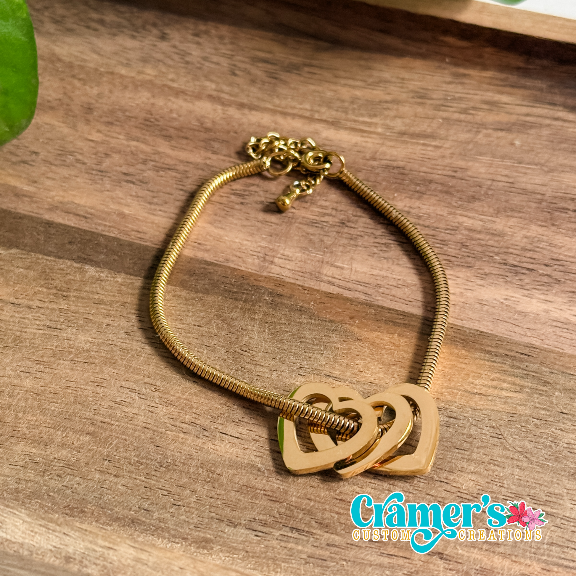 Gold bracelet with heart designs on a wooden surface, featuring logo.