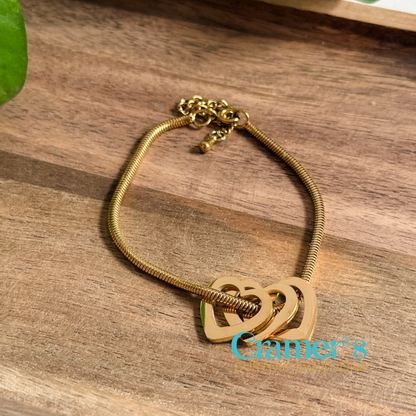 Gold bracelet with heart designs on a wooden surface, featuring logo.