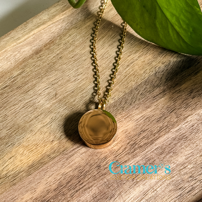 A gold-colored circle urn pendant necklace with the initials 'BLE' engraved on the front, displayed on a wooden surface.