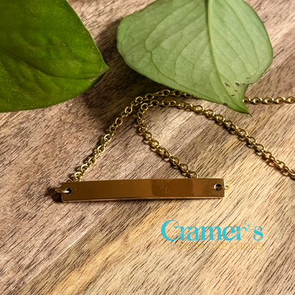 A gold-colored horizontal bar bracelet attached to a chain, displayed on a wooden surface with a large green leaf in the background.