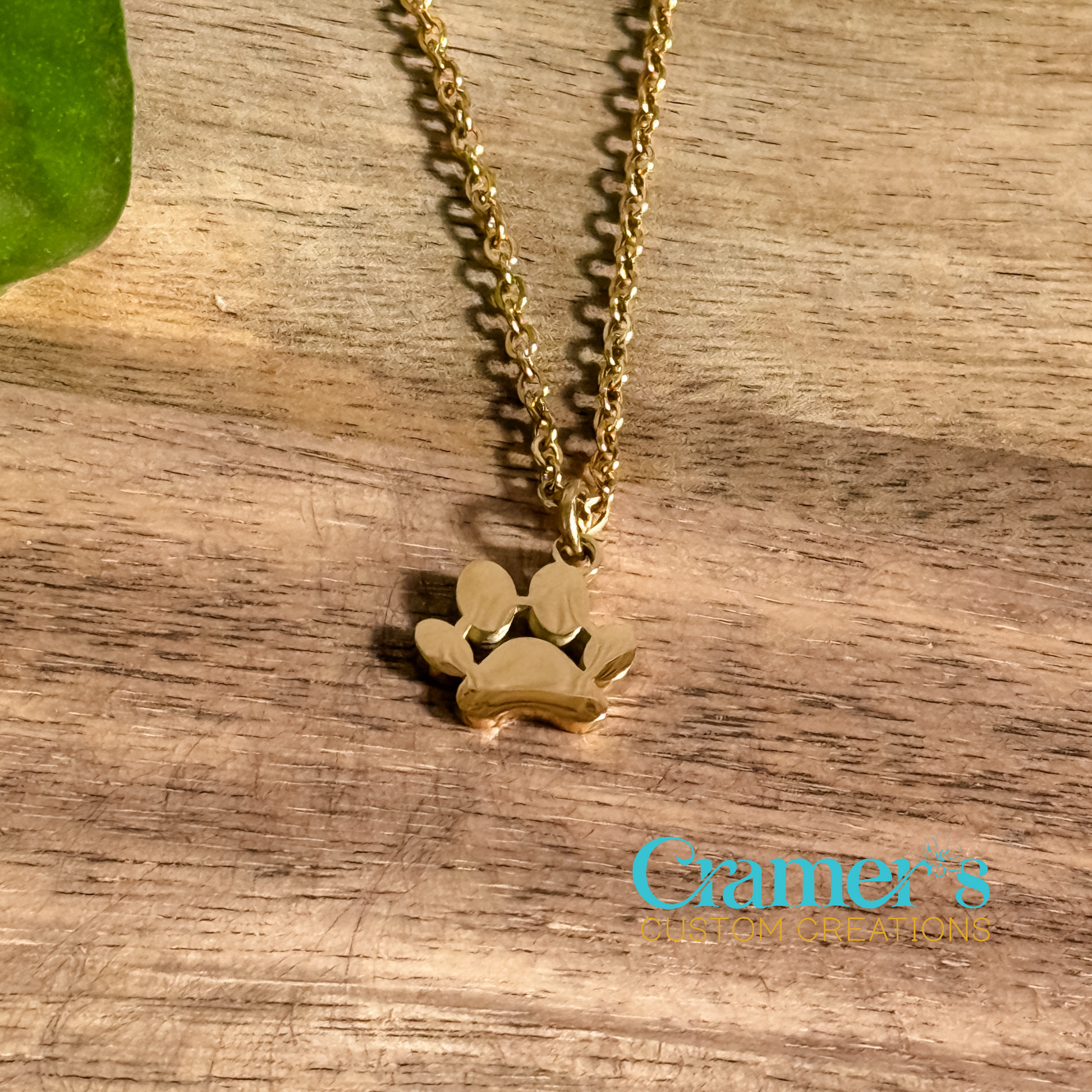A gold-colored paw print pendant necklace on a wooden surface, with a chain and Cramer's Custom Creations logo in the background.