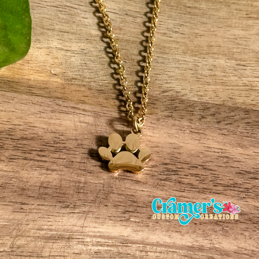 A gold-colored paw print pendant necklace on a wooden surface, with a chain and Cramer's Custom Creations logo in the background.