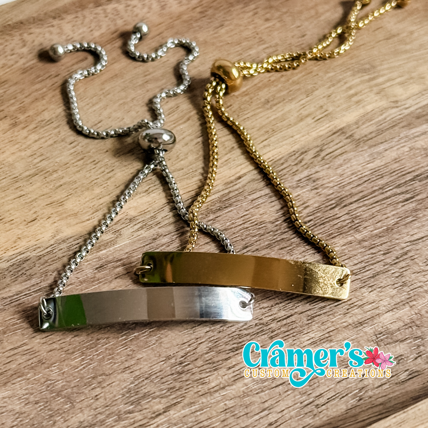 Two bracelets, one gold and one silver, on a wooden surface with 'Cramer's Custom Creations' branding.