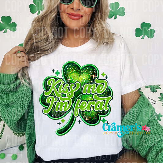 girl wearing a white tee with kiss my im feral on it and a faux embroidered shamrock, theres a st pattys day background