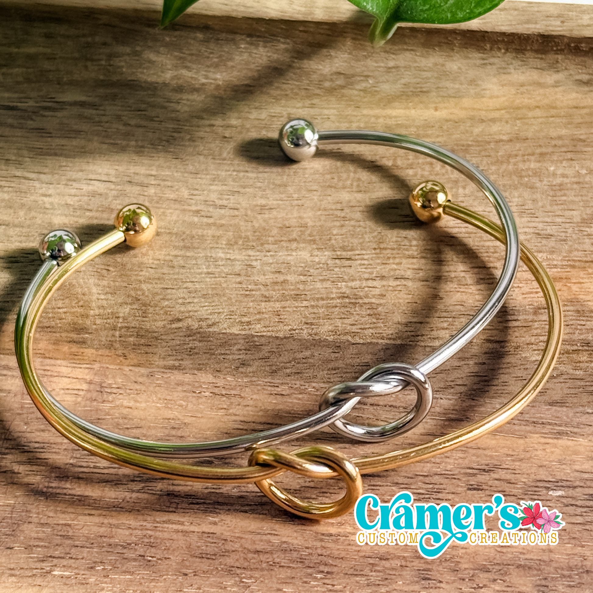 A knotted charm bracelet with a silver and gold color scheme, featuring a simple loop knot design and small metal balls on each end, displayed on a wooden surface.