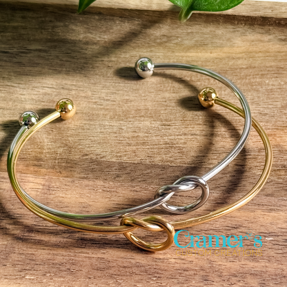 A knotted charm bracelet with a silver and gold color scheme, featuring a simple loop knot design and small metal balls on each end, displayed on a wooden surface.