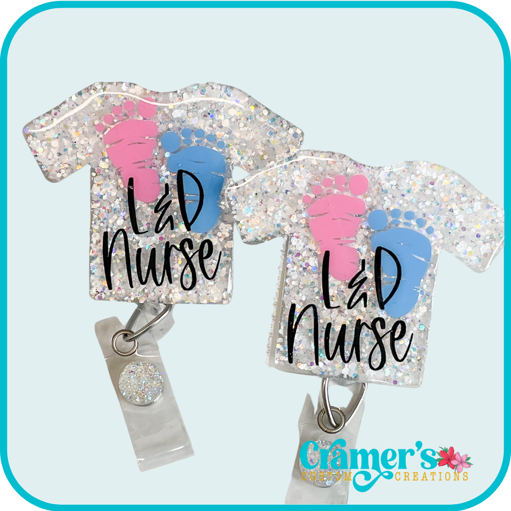 badge shaped like a scrub top with white glitter, a pink and a blue foorprint, and the words 'L&D Nurse'