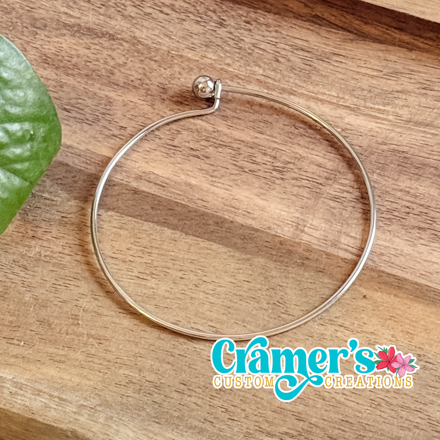 Silver minimalist bangle bracelet on a wooden surface with 'Cramer's Custom Creations' logo.