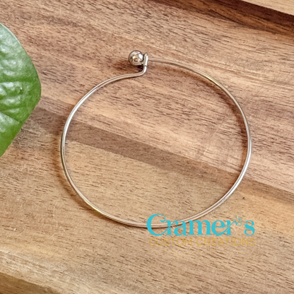 Silver minimalist bangle bracelet on a wooden surface with 'Cramer's Custom Creations' logo.