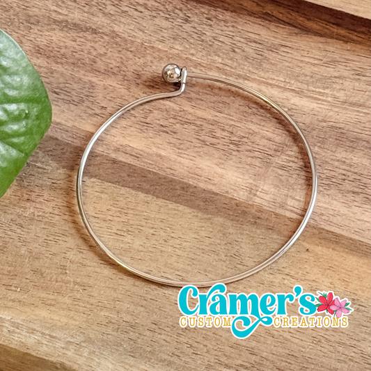 Silver minimalist bangle bracelet on a wooden surface with 'Cramer's Custom Creations' logo.