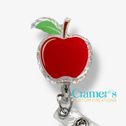 Red apple-shaped badge holder with a green leaf on a white background, featuring the brand 'Cramer's Custom Creations'.