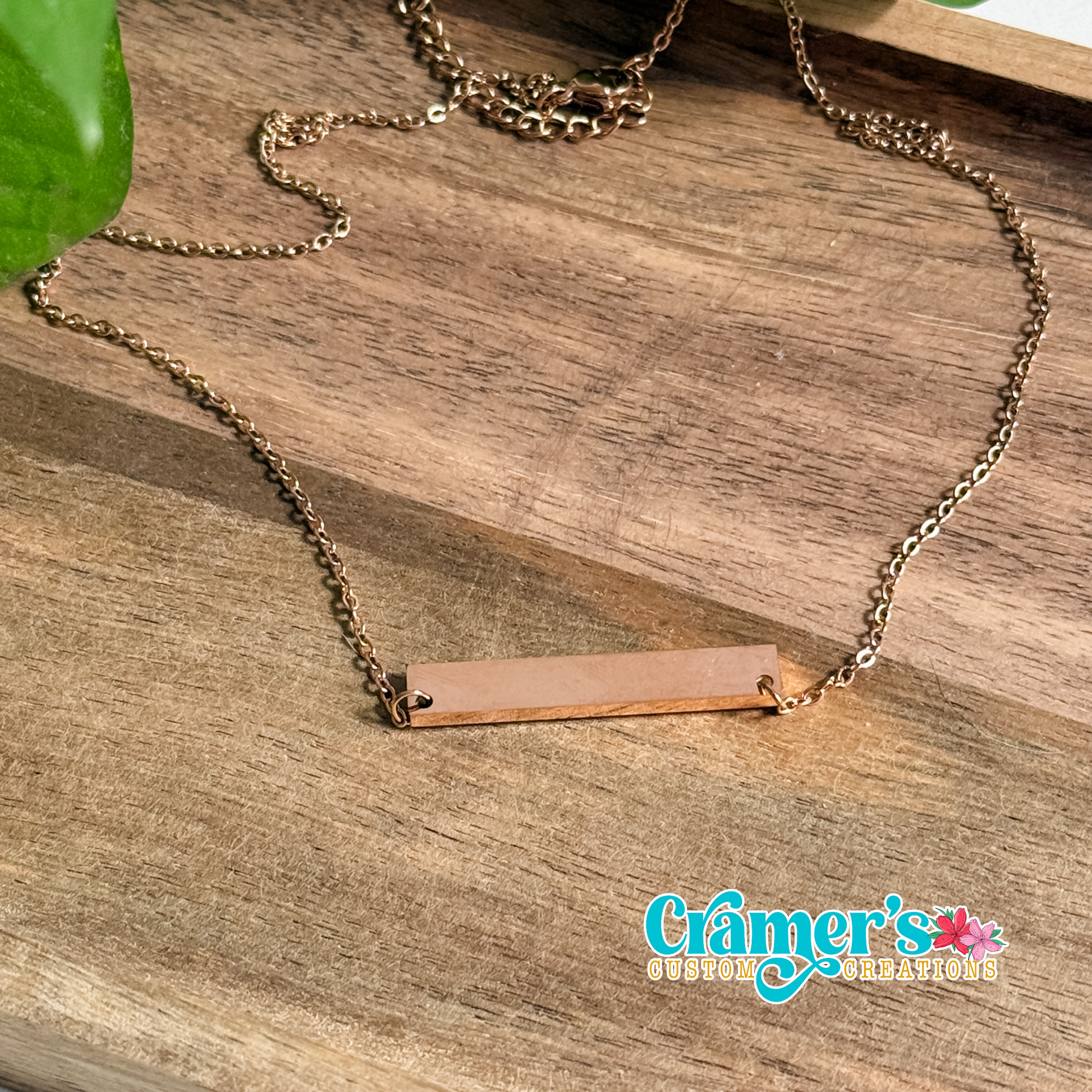 Rose gold necklace with a rectangular pendant on a wooden surface, featuring the brand logo