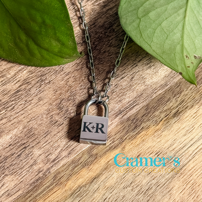 A petite lock pendant necklace with the initials 'K-R' on it, displayed on a wooden surface with green leaves in the background.