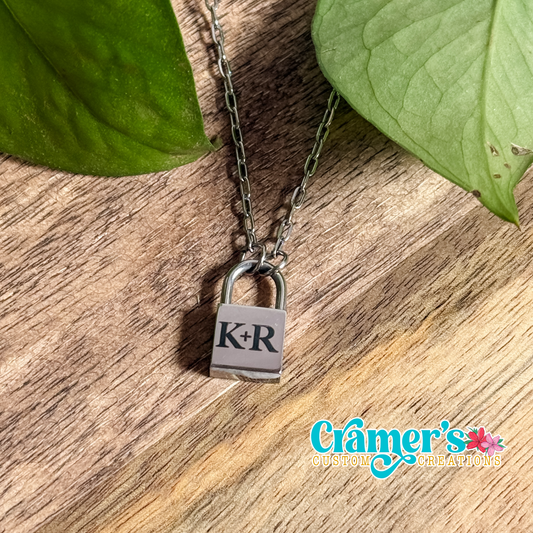 A petite lock pendant necklace with the initials 'K-R' on it, displayed on a wooden surface with green leaves in the background.