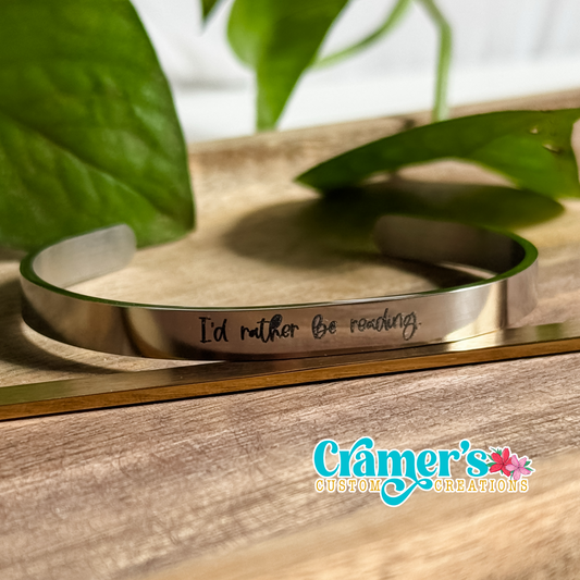 A silver bangle bracelet with the inscription 'I'd rather be reading' on one side, displayed on a wooden surface with leaves in the background.