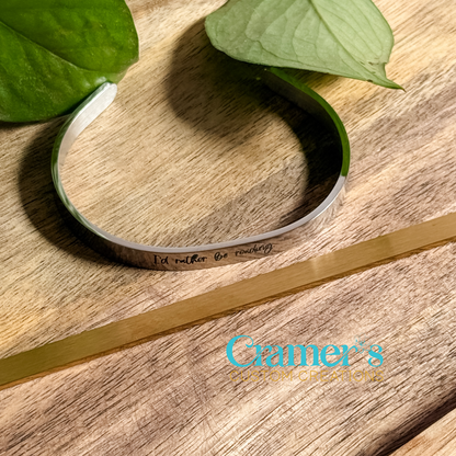 a silver bangle bracelet with writing on it, laying next to a gold blank one, on a wood background