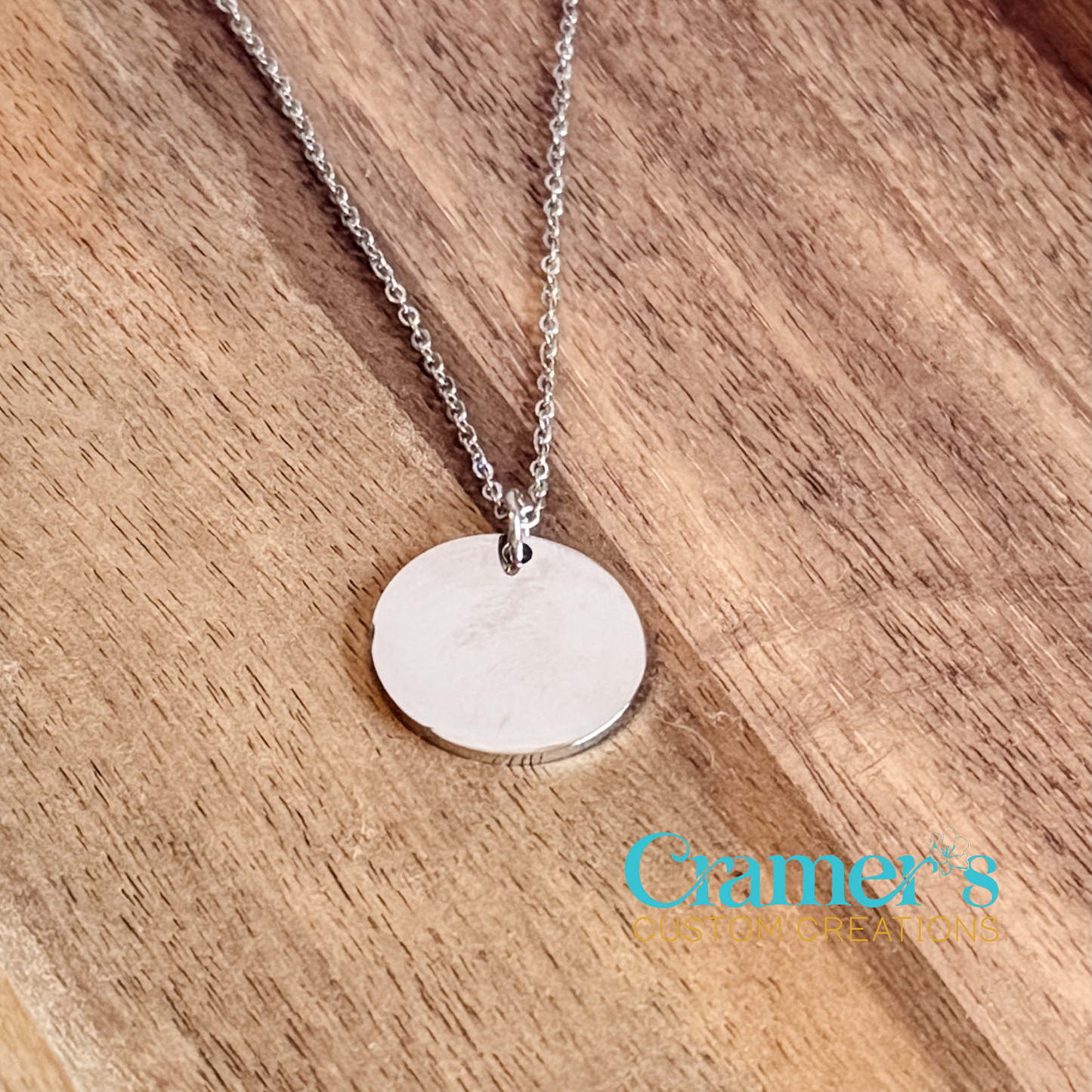 Silver necklace with a 20 mm round pendant on a wooden surface, featuring the 'Cramer's Custom Creations' logo.
