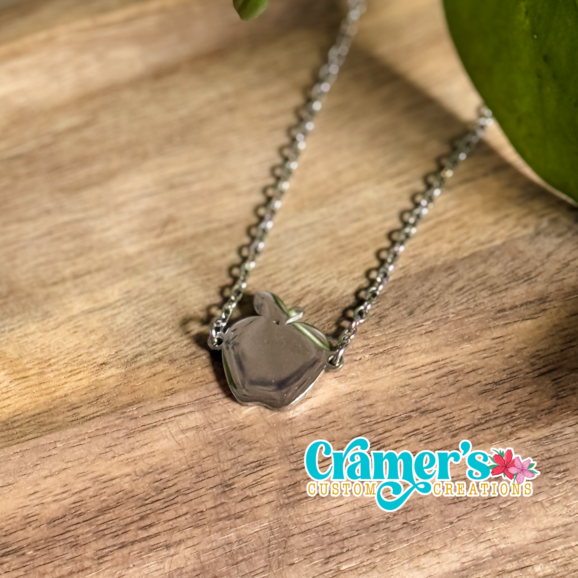 A silver necklace with a silver apple pendant on a wooden surface, with the brand mark 'Cramer's Custom Creations' visible.