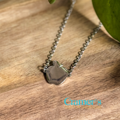A silver necklace with a silver apple pendant on a wooden surface, with the brand mark 'Cramer's Custom Creations' visible.