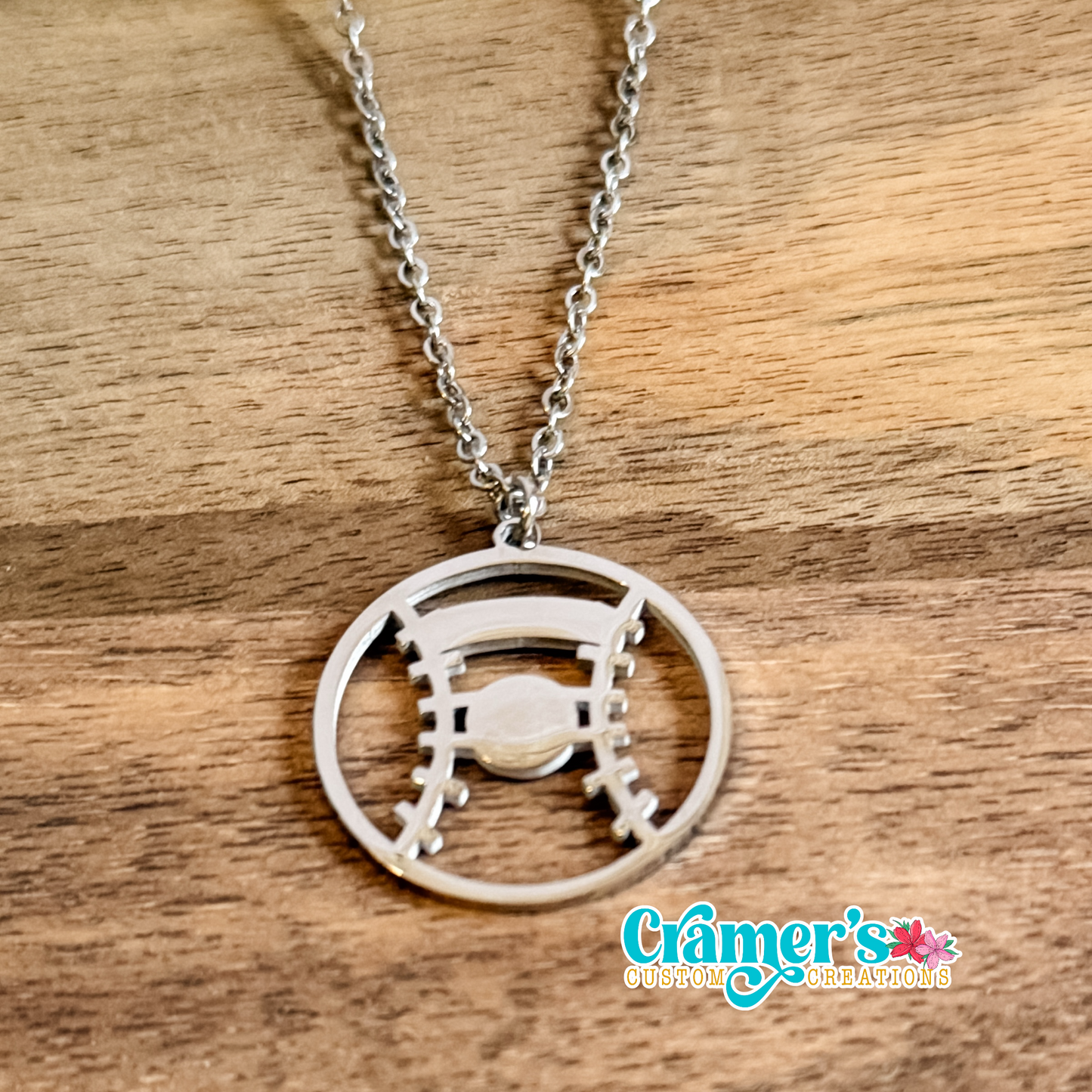 Silver necklace with a baseball or softball  pendant on a wooden surface, featuring the Cramer's Custom Jewelry logo.