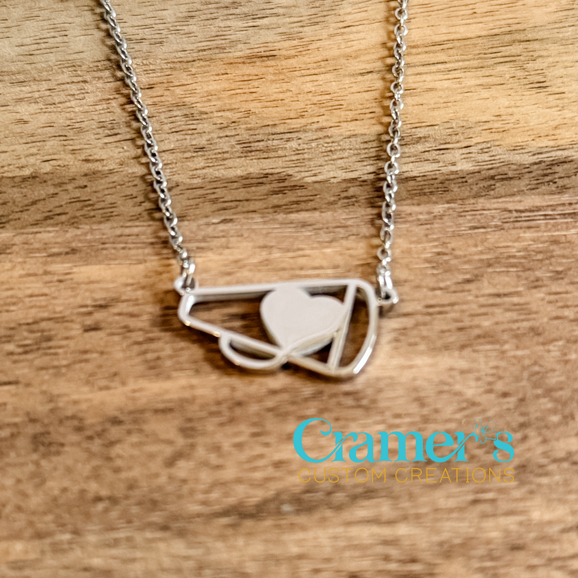 Silver necklace with a cheer megaphone pendant on a wooden surface, featuring Cramer's Custom Creations logo.