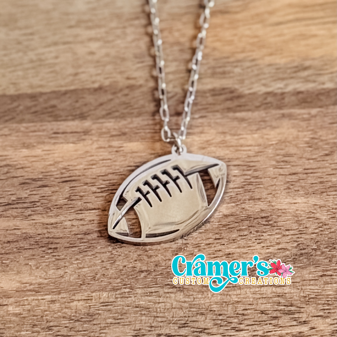 Silver football-shaped pendant necklace on a wooden surface with 'Gramer's Custom Creations' branding.