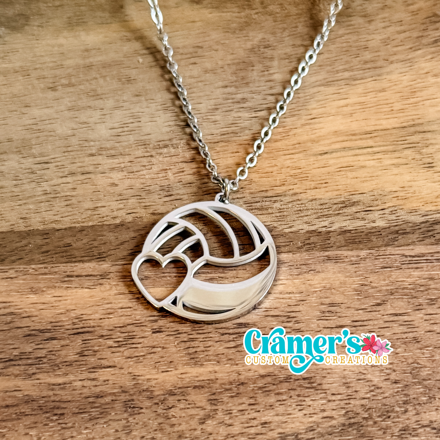Silver necklace with a volleyball pendant on a wooden surface, featuring Cramer's Custom Creations logo.