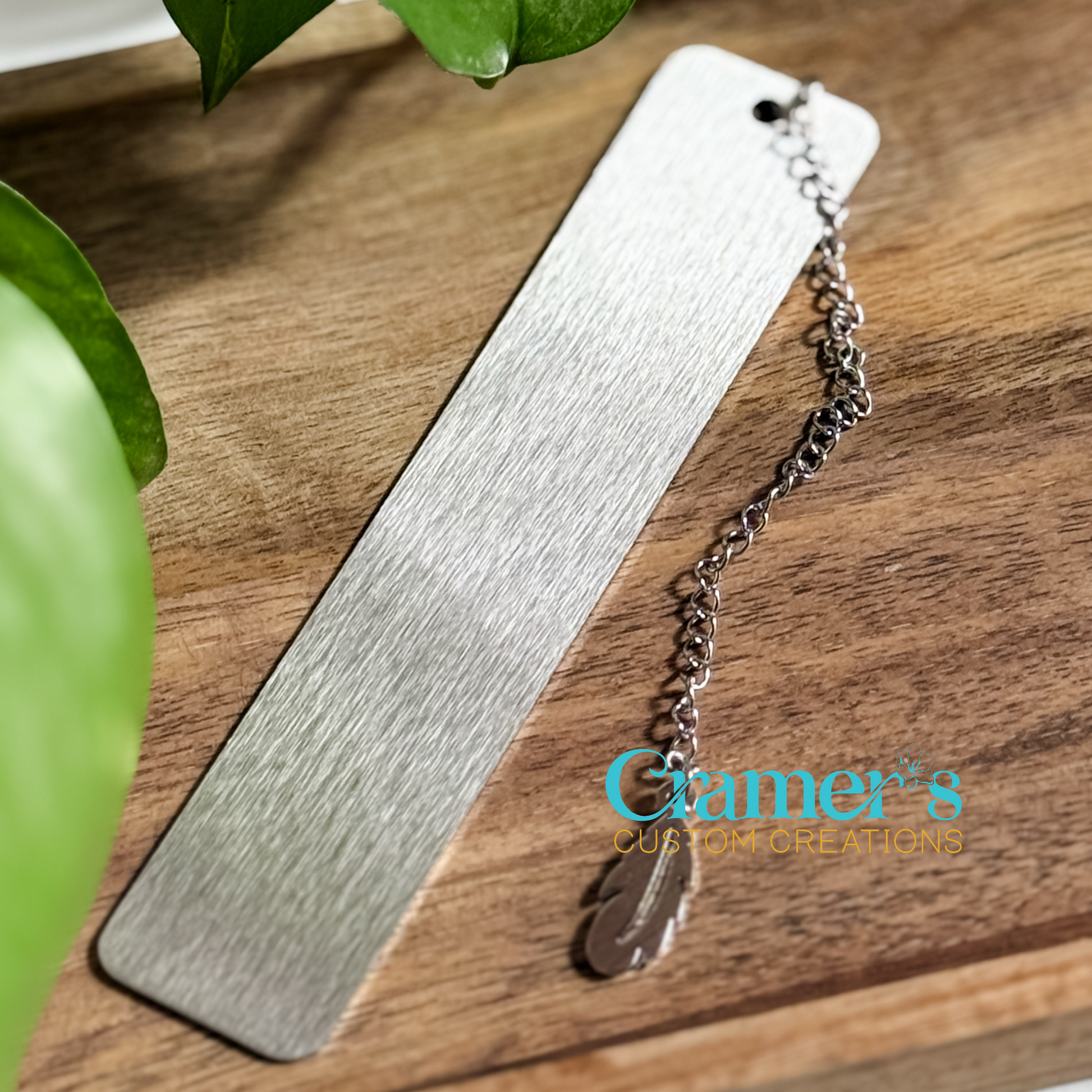 A silver metal bookmark with a chain tassel resting on a wooden surface, with a leafy background.