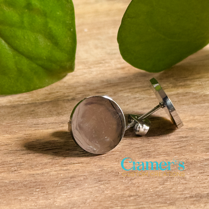 A pair of stainless steel circle stud earrings with a blank engraving surface on a wooden surface with a leaf in the background.