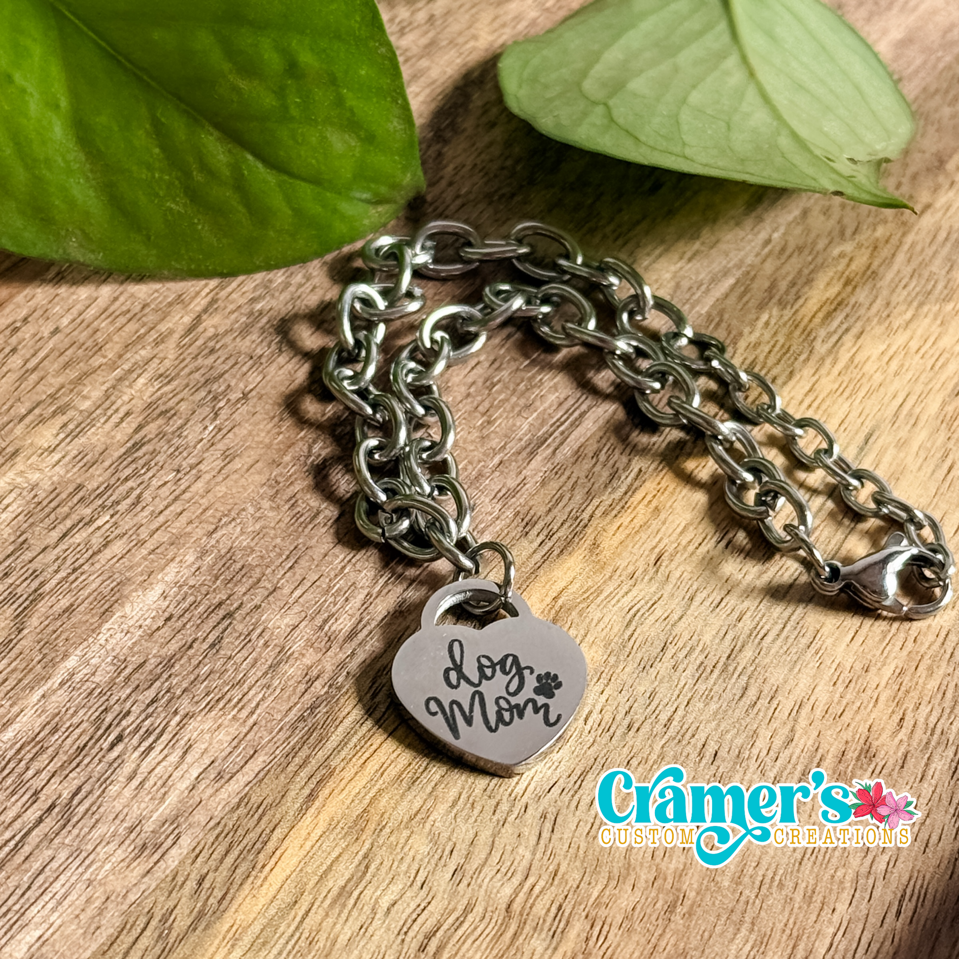 A silver heart pendant bracelet with the inscription 'Love Mom' on a wooden surface, accompanied by leaf accents in the background.