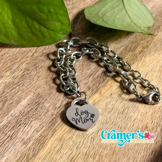 A silver heart pendant bracelet with the inscription 'Love Mom' on a wooden surface, accompanied by leaf accents in the background.