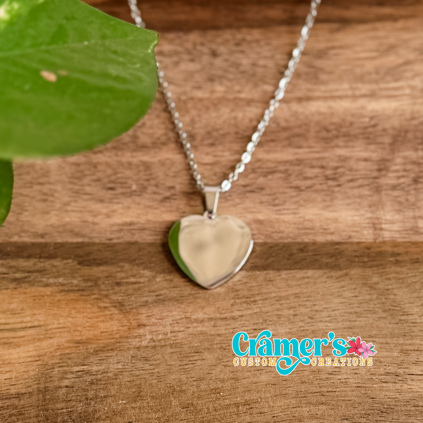 Heart-shaped silver pendant necklace on a wooden surface with a brand logo in the corner.