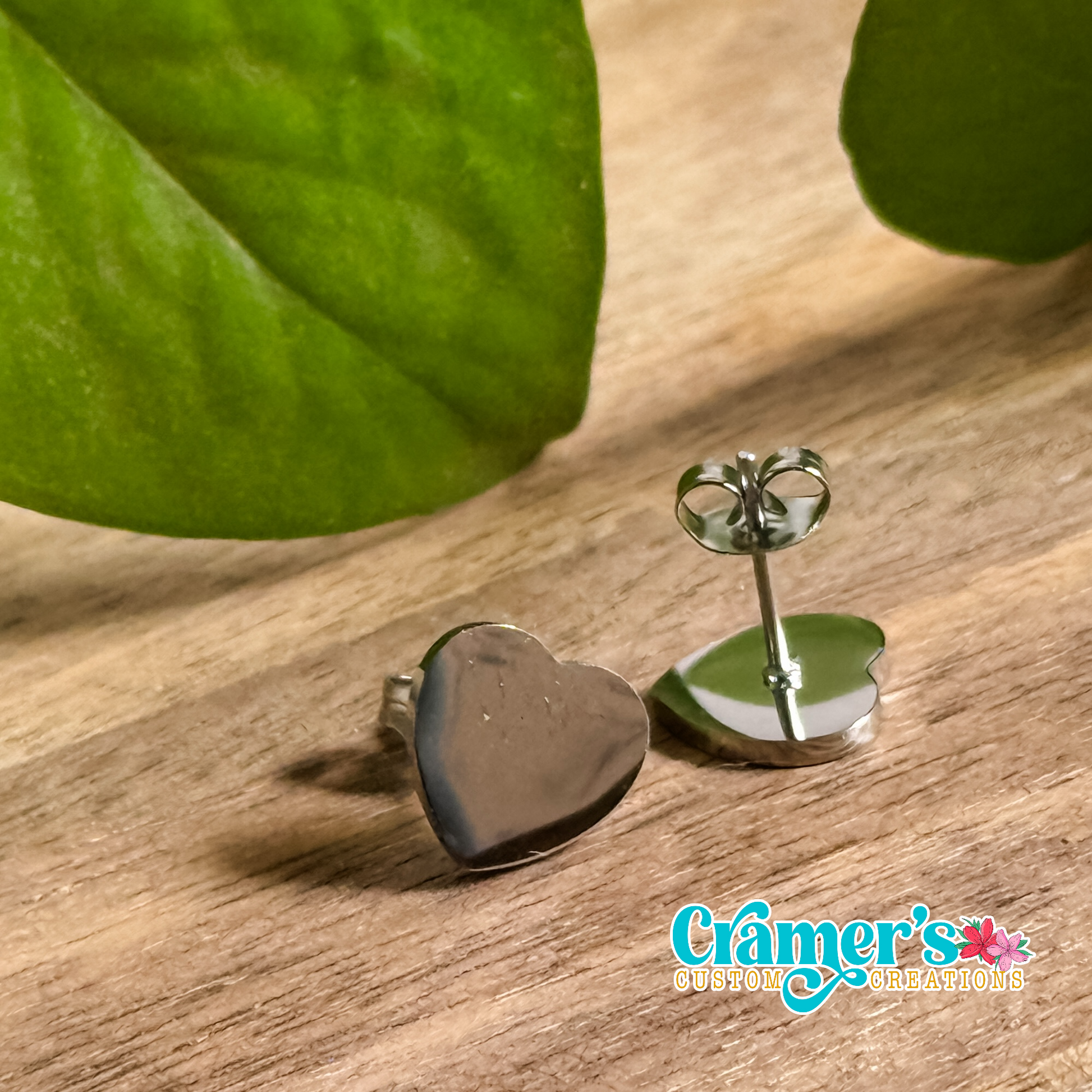 A pair of heart-shaped stud earrings in a silver color, displayed on a wooden surface with a leaf in the background.