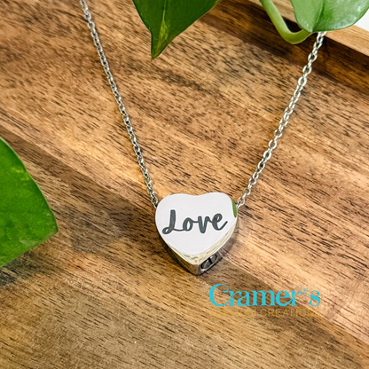 A heart-shaped urn pendant necklace with the word 'Love' inscribed on it, displayed on a wooden surface with green leaves in the background.
