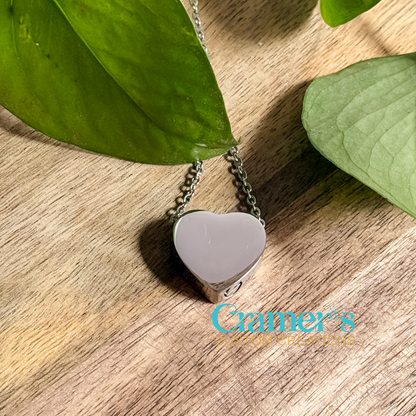 A silver heart-shaped urn pendant necklace made of stainless steel, displayed on a wooden surface with green leaves in the background. 