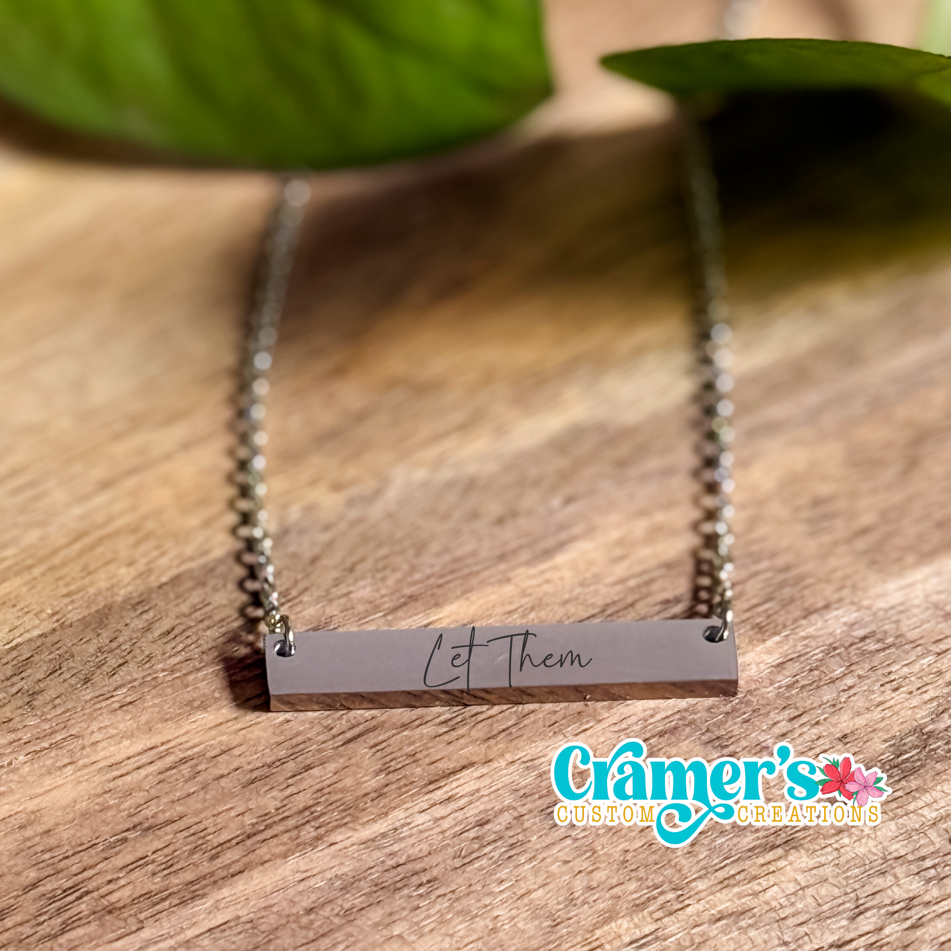 A horizontal bar necklace with the inscription 'Let them' written in cursive on a chain, placed on a wooden surface with a leaf in the background.