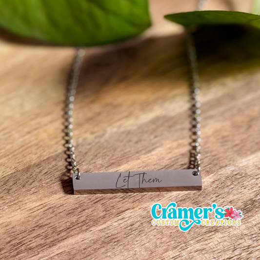 A horizontal bar necklace with the inscription 'Let them' written in cursive on a chain, placed on a wooden surface with a leaf in the background.