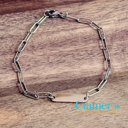 Silver chain bracelet with a nameplate on a wooden surface, featuring 'Cramer's Custom Creations' logo.